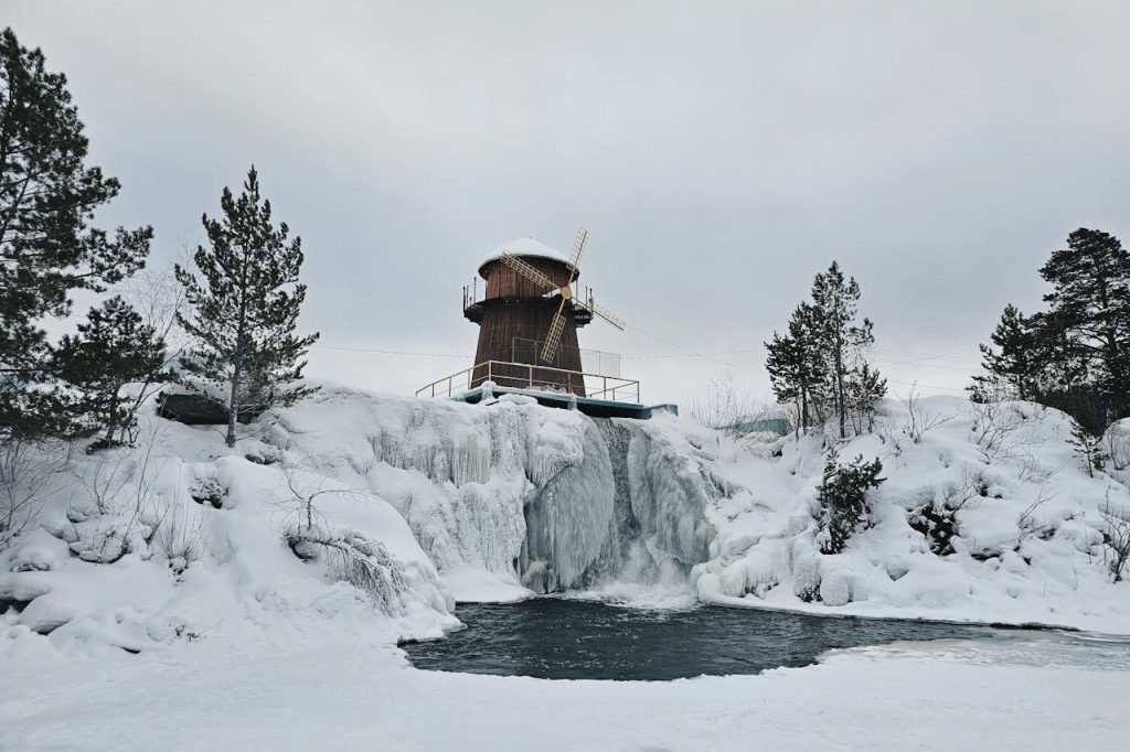 A picturesque wooden windmill on a snowy hill with a frozen waterfall and surrounding trees.