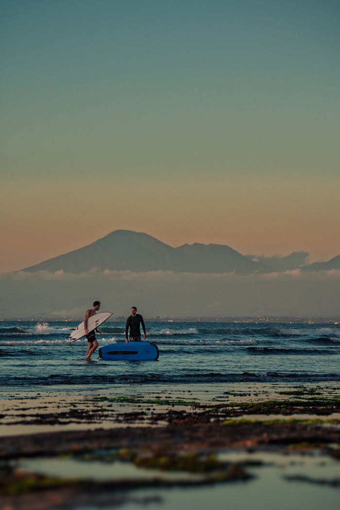 Two surfers at shoreline with surfboards against scenic mountain backdrop during sunset.